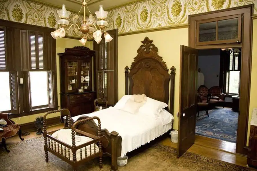 A vintage bedroom with ornate wooden furniture including a bed, baby crib, wardrobe, chandelier, and patterned wallpaper