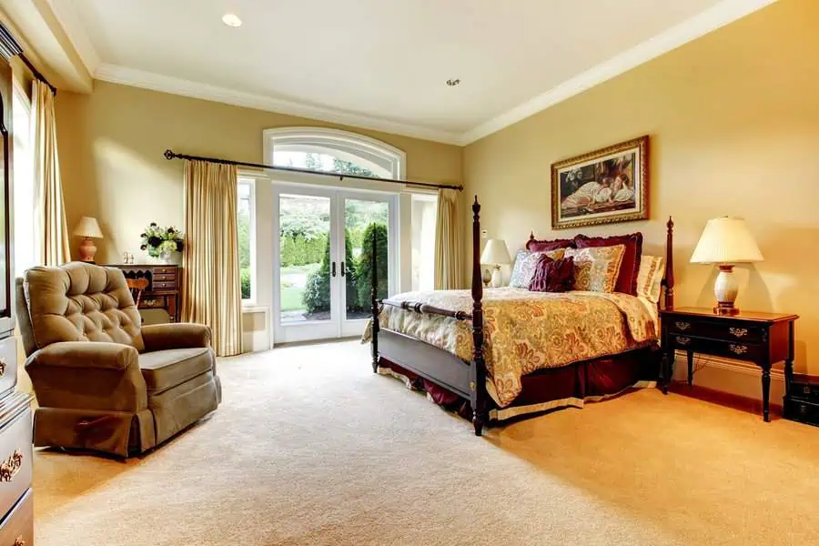 Warm, elegant bedroom with large bed, armchair, side tables with lamps, dresser, and French doors opening to a garden