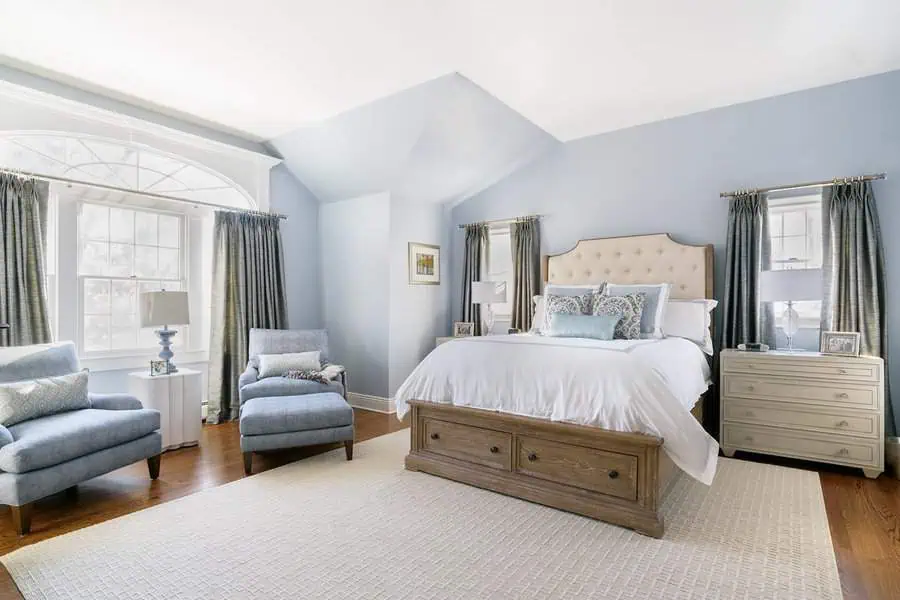 Bedroom with light blue walls, large bed, armchairs, and a big window with curtains