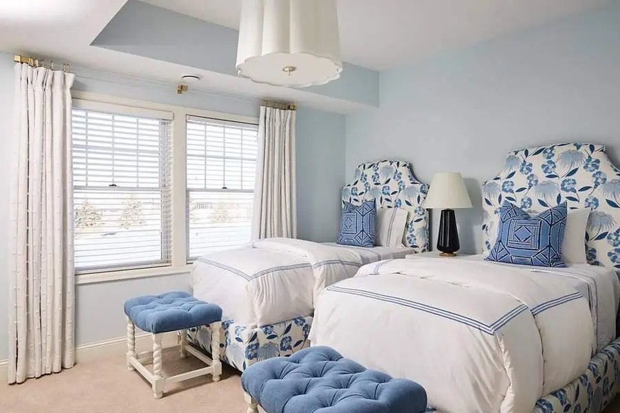 Two twin beds in a bright blue and white bedroom with matching stools and a window view