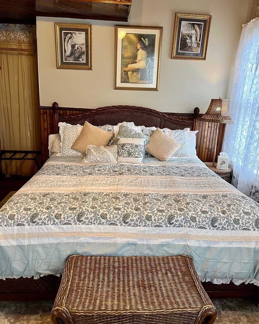 A cozy bedroom with a large bed, decorative pillows, framed artwork, wicker bench, and a lamp next to a sheer-curtained window