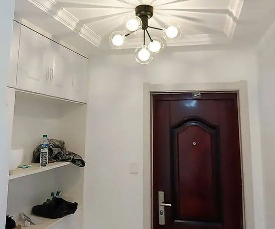 A well-lit hallway with a modern ceiling light fixture, red wooden door, and a white shelf