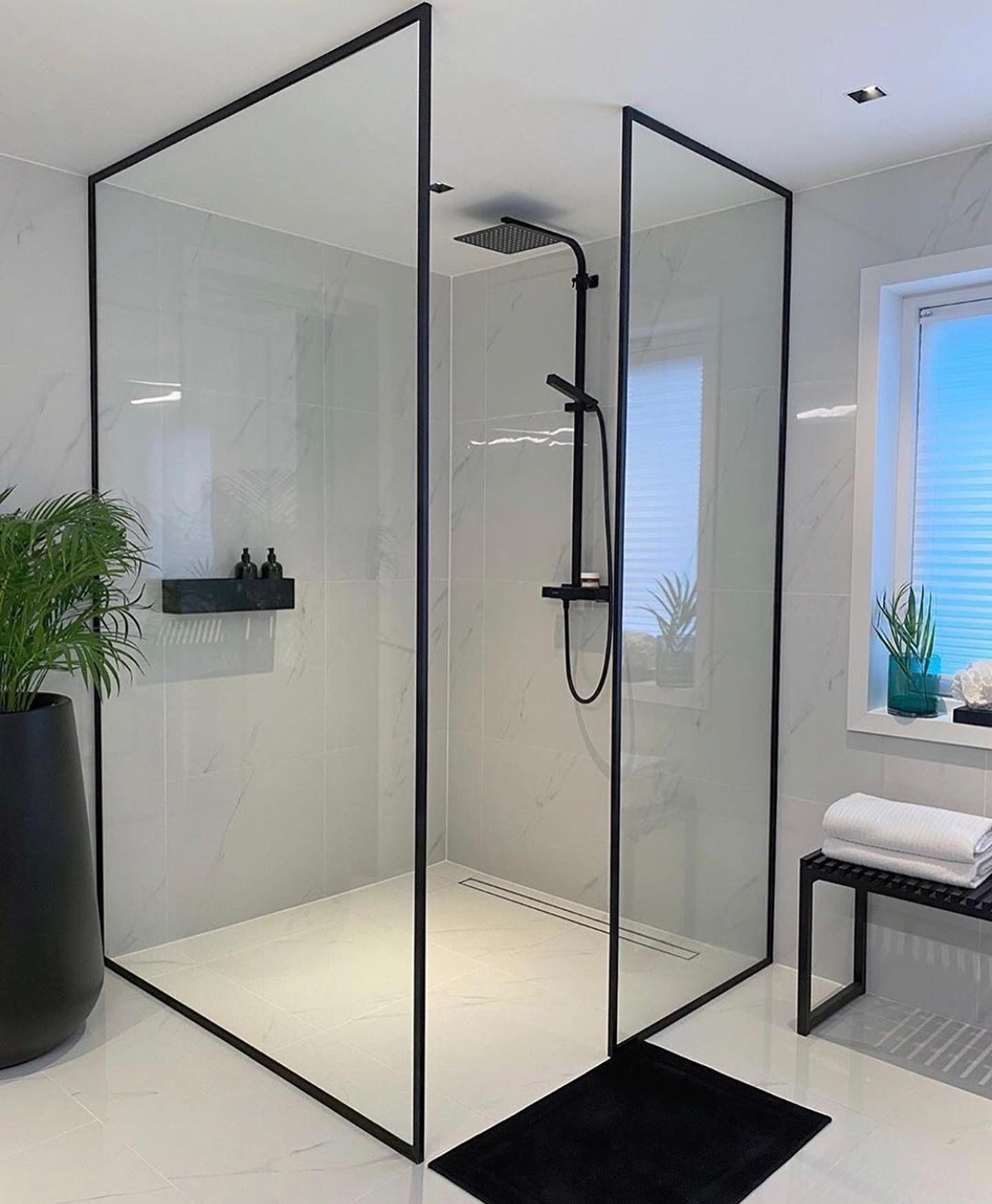 Modern minimalist design shower with black trim, glass panels, plants, and a black mat on the white floor