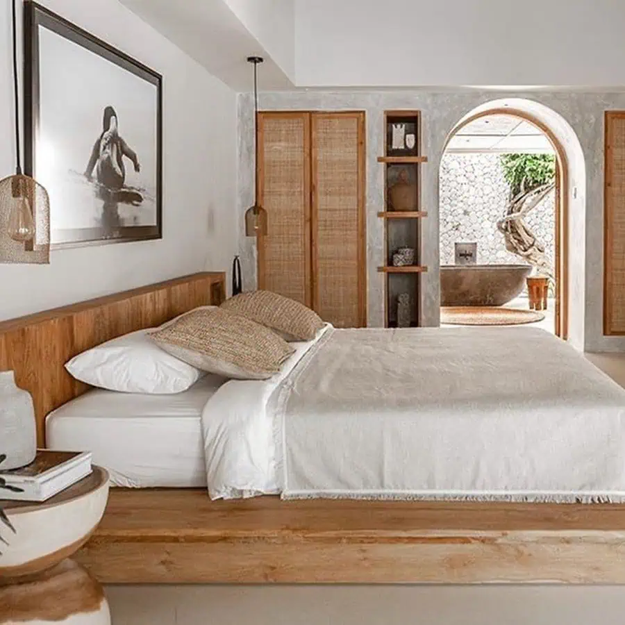 Minimalist bedroom featuring a wooden bed with white bedding, art on the wall, wicker lamps, and an open archway leading to a bathroom