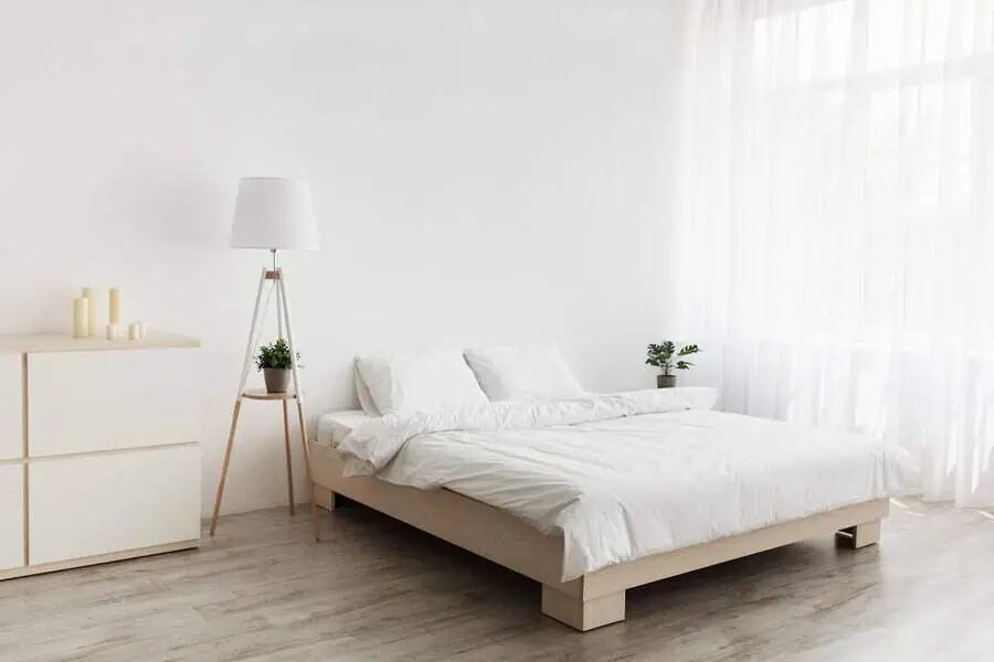 Minimalist bedroom with a low wooden bed, white bedding, a dresser, and a floor lamp near a large window.