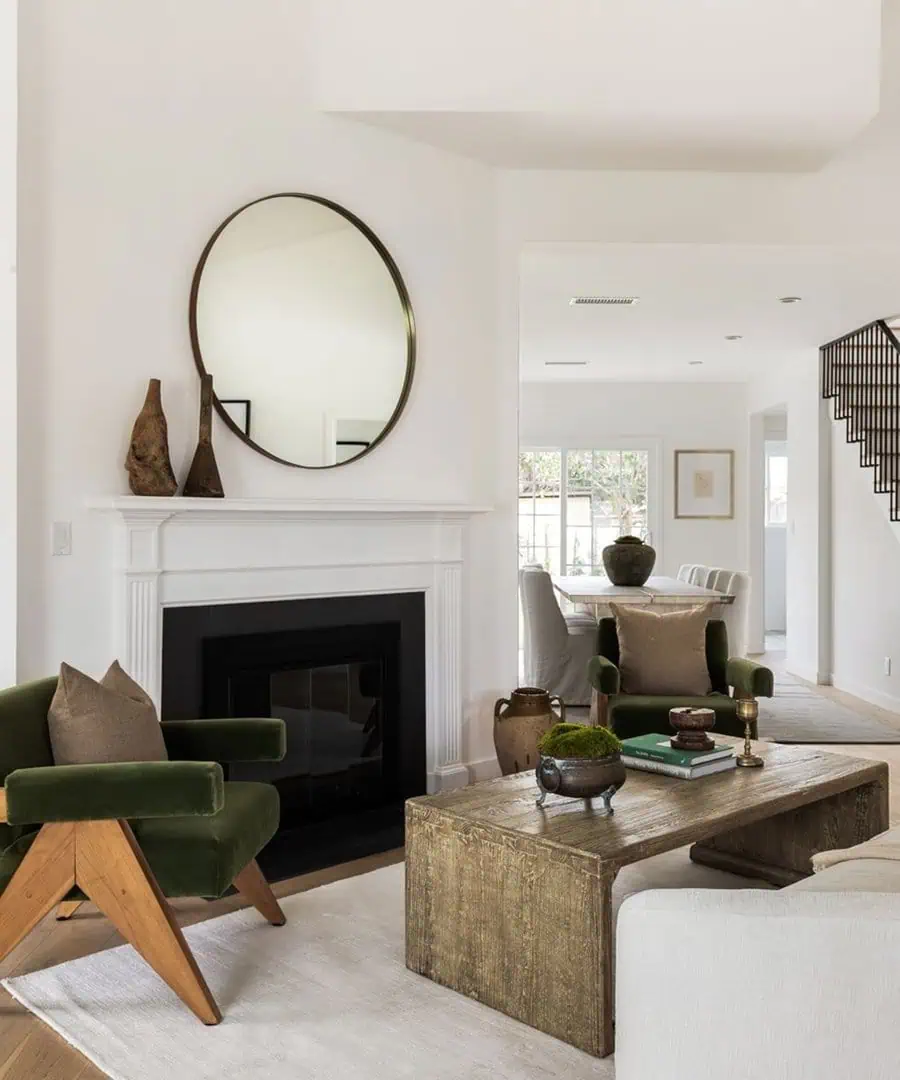 Elegant living room with green velvet chairs, a rustic wooden coffee table, a round mirror over the fireplace, and neutral decor