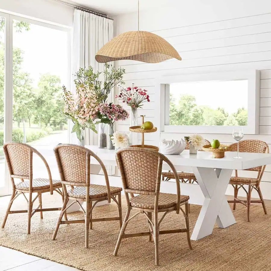 Bright dining area featuring a white table, wicker chairs, floral centerpiece, large window, and a woven pendant light