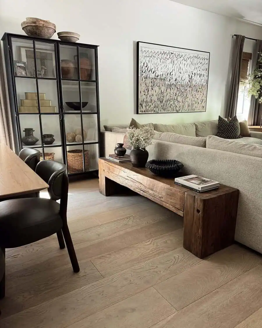 Modern living room with neutral tones, featuring a glass cabinet, abstract wall art, a wooden table, and a beige sectional sofa