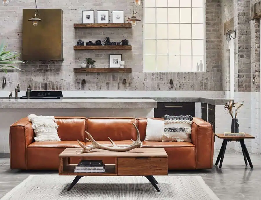 A stylish loft with a brown leather sofa, rustic coffee table, and open kitchen shelving next to a large window