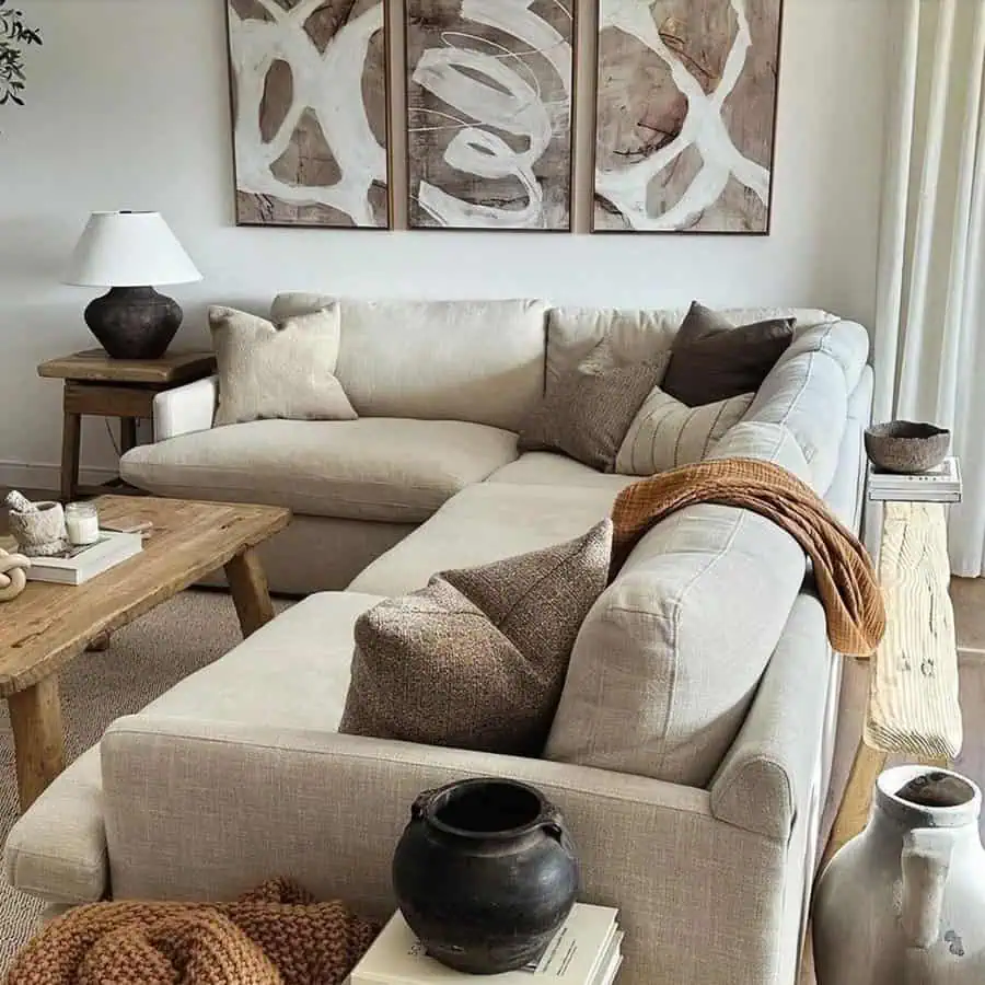 A cozy, neutral-toned living room with a beige sectional sofa, wooden tables, abstract wall art, and decorative vases