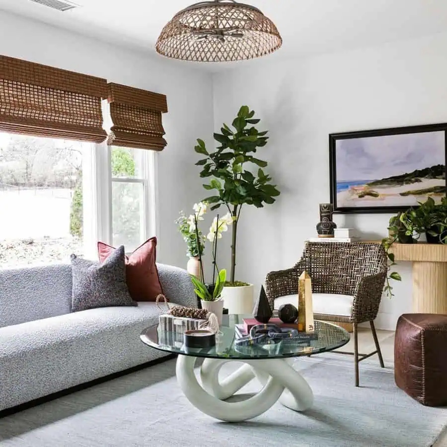 Modern living room with a gray sofa, wicker chair, glass coffee table, large plant, and wall art of a beach scene