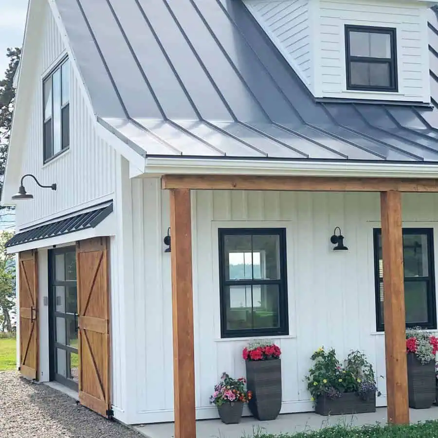 A white modern farmhouse with a metal roof, black windows, wooden accents, and potted flowers under the porch