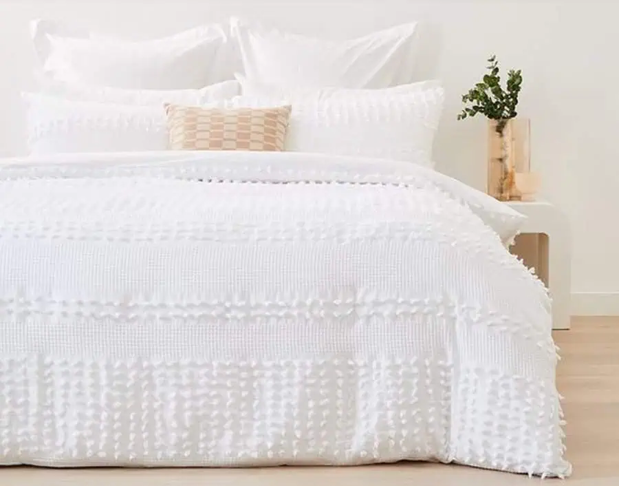 A bed with white textured bedding, pillows, and a small beige pillow; a nightstand with plants.