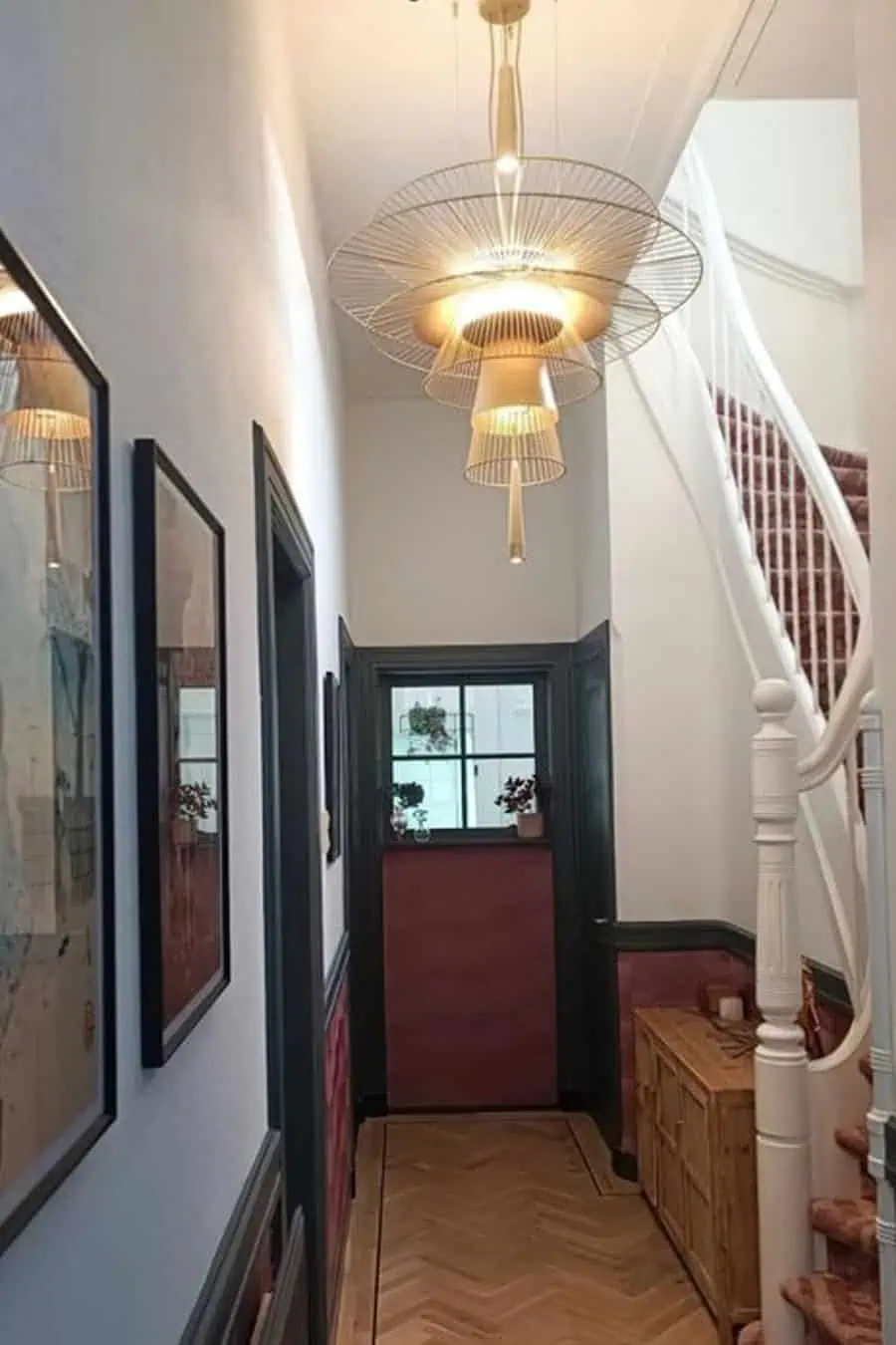 A narrow hallway with framed pictures on the wall, a stylish pendant light, wooden furniture, and a staircase