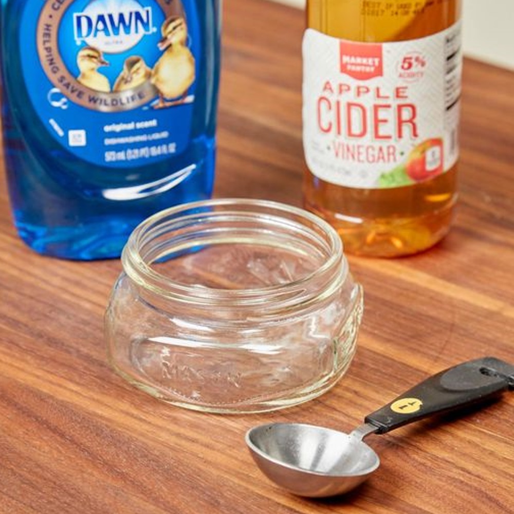 Dish soap, apple cider vinegar, an empty jar, and a measuring spoon on a table