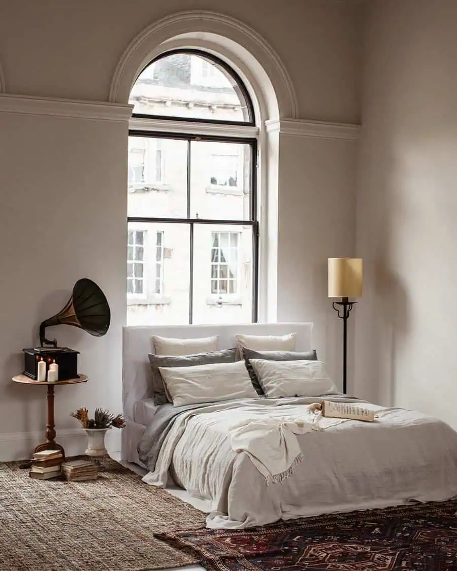 Cozy bedroom with a large arched window, soft bedding, vintage decor, and a gramophone on a side table.