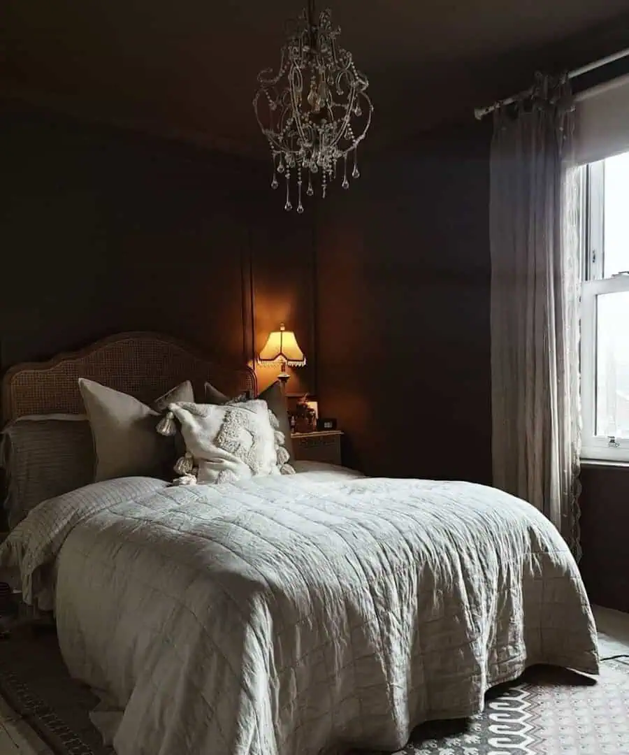 A cozy bedroom with a soft-lit lamp, an ornate chandelier, and a large window with white curtains beside a neatly made bed