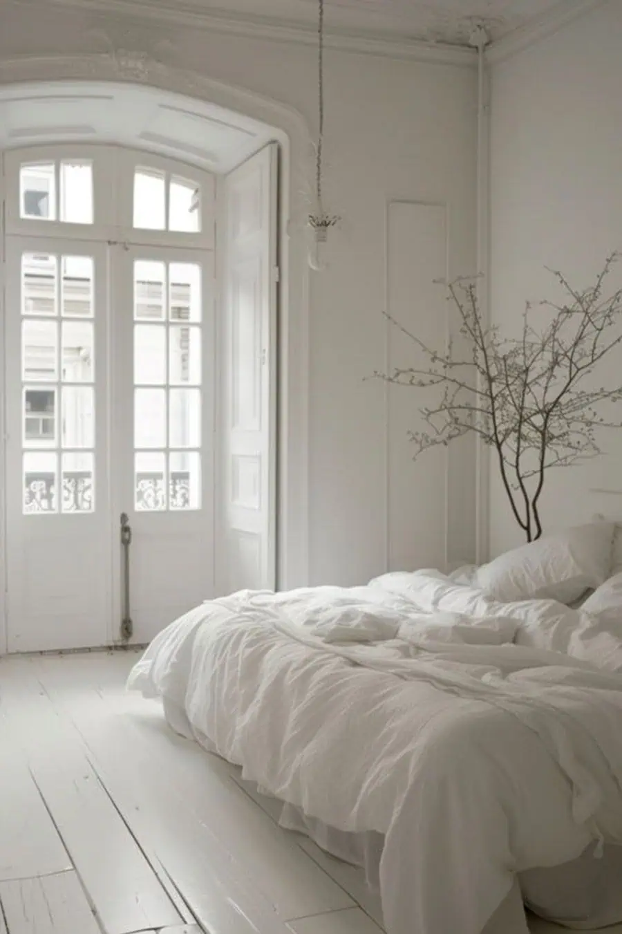 Serene bedroom with white bedding, French doors, and minimalist decor featuring a delicate branch arrangement.