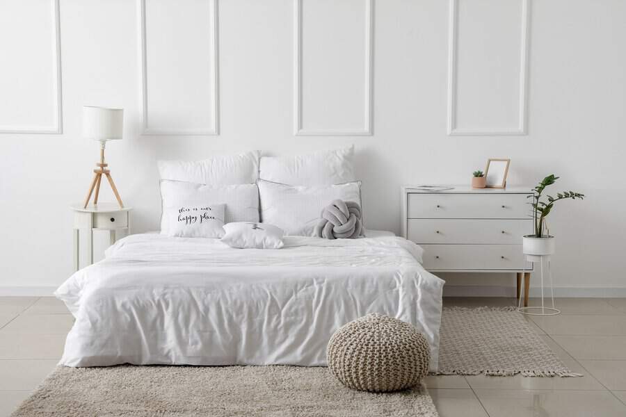 Cozy bedroom with a white bed, decorative pillows, a knitted pouf, and light wooden furniture.