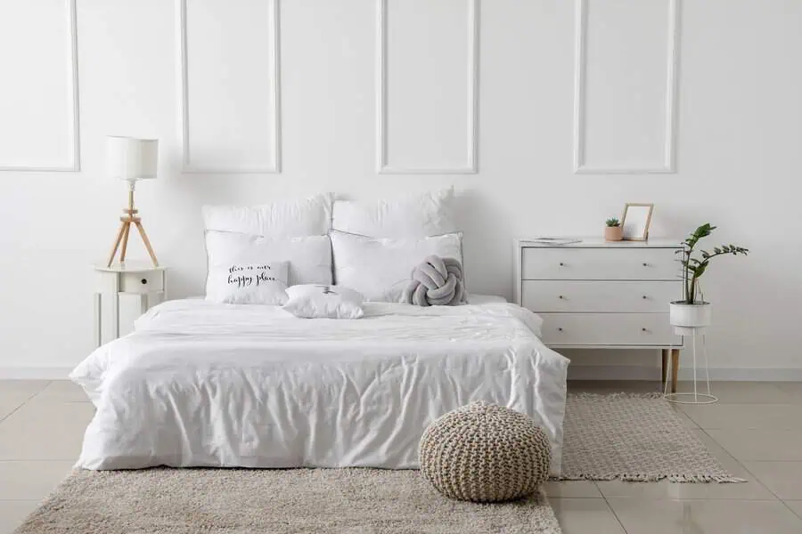 Cozy bedroom with a white bed, decorative pillows, a knitted pouf, and light wooden furniture.