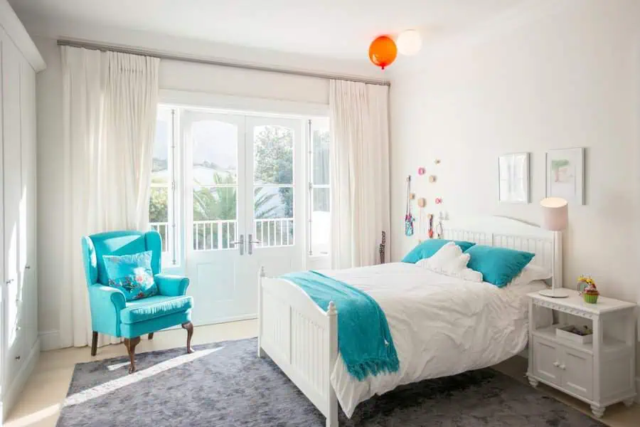 Bright bedroom with a white bed, teal accents, a cozy armchair, and large windows opening to a balcony.