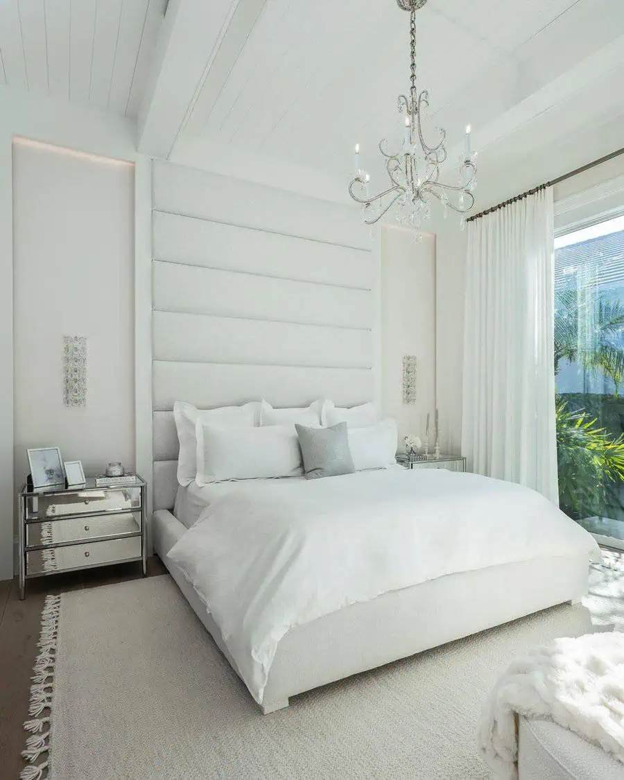 Elegant white bedroom with plush bed, chandelier, mirrored nightstands, and large window with sheer curtains.