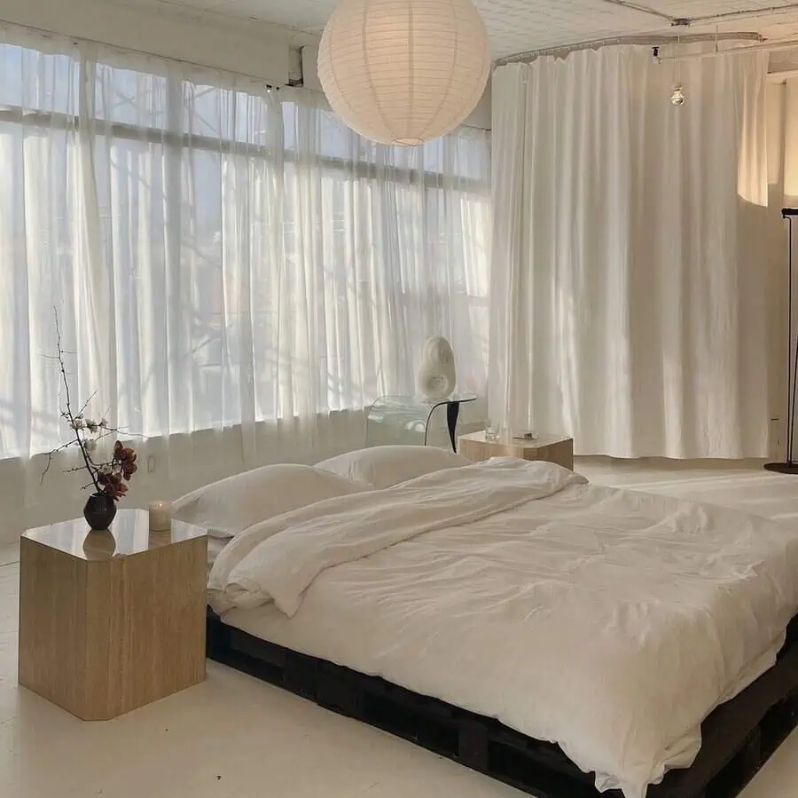 Minimalist bedroom with a low pallet bed, sheer curtains, soft bedding, and natural light filtering through large windows.