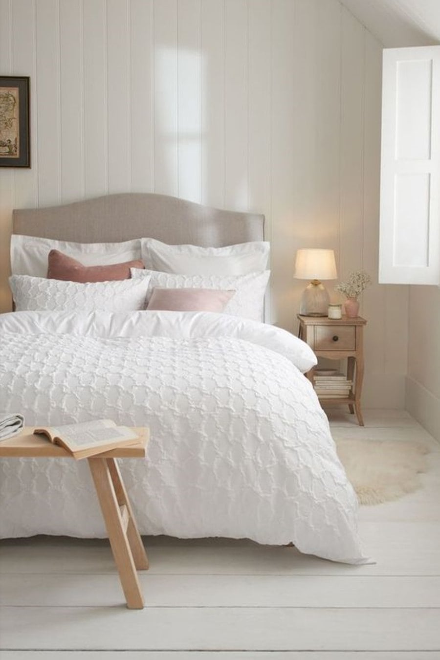 Cozy bedroom with white bedding, bedside table, lamp, window, and a book on a wooden bench.