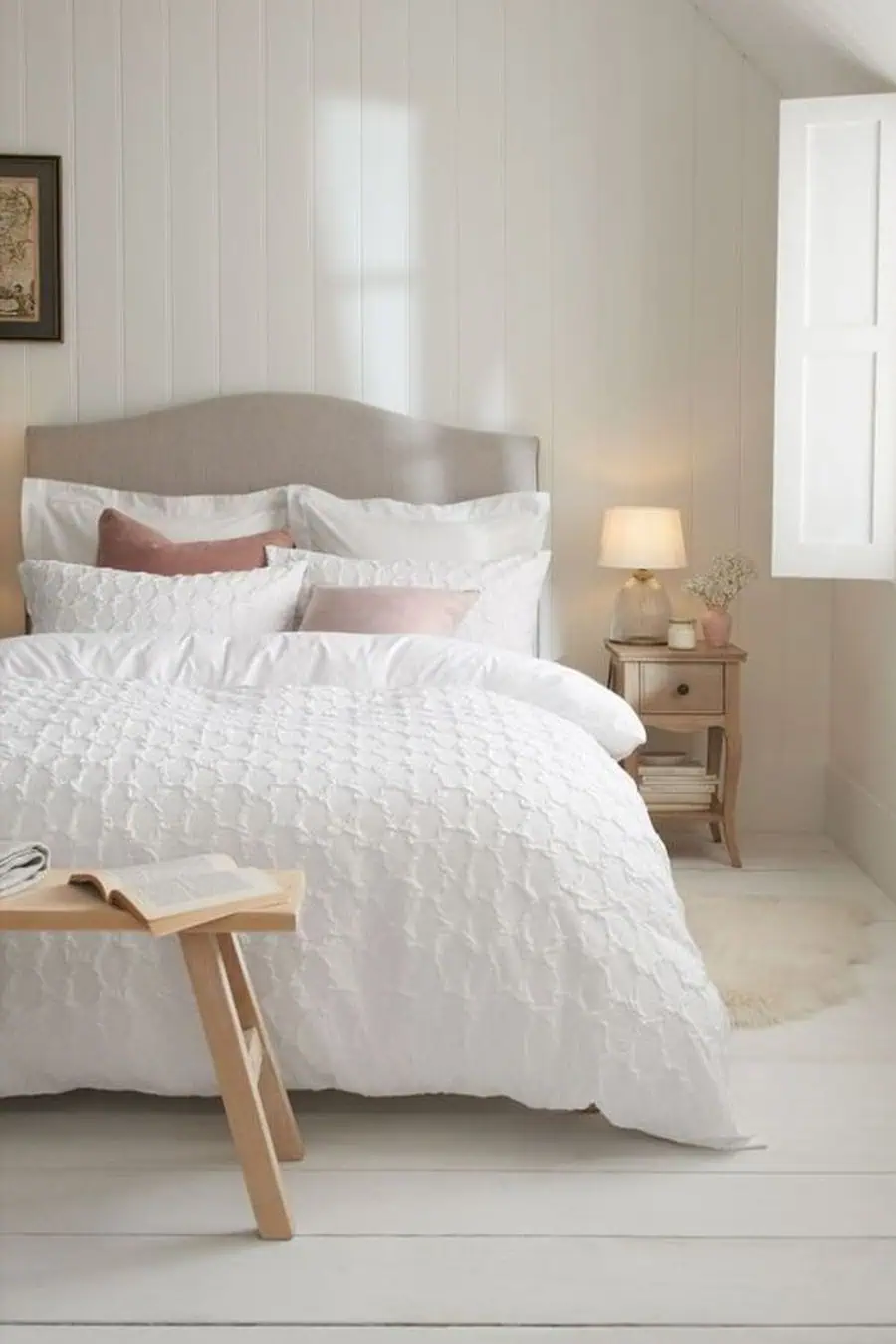 Cozy bedroom with white bedding, bedside table, lamp, window, and a book on a wooden bench.