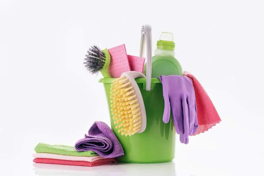 A green bucket with various cleaning supplies including brushes, sponges, a bottle, gloves, and towels against a white background