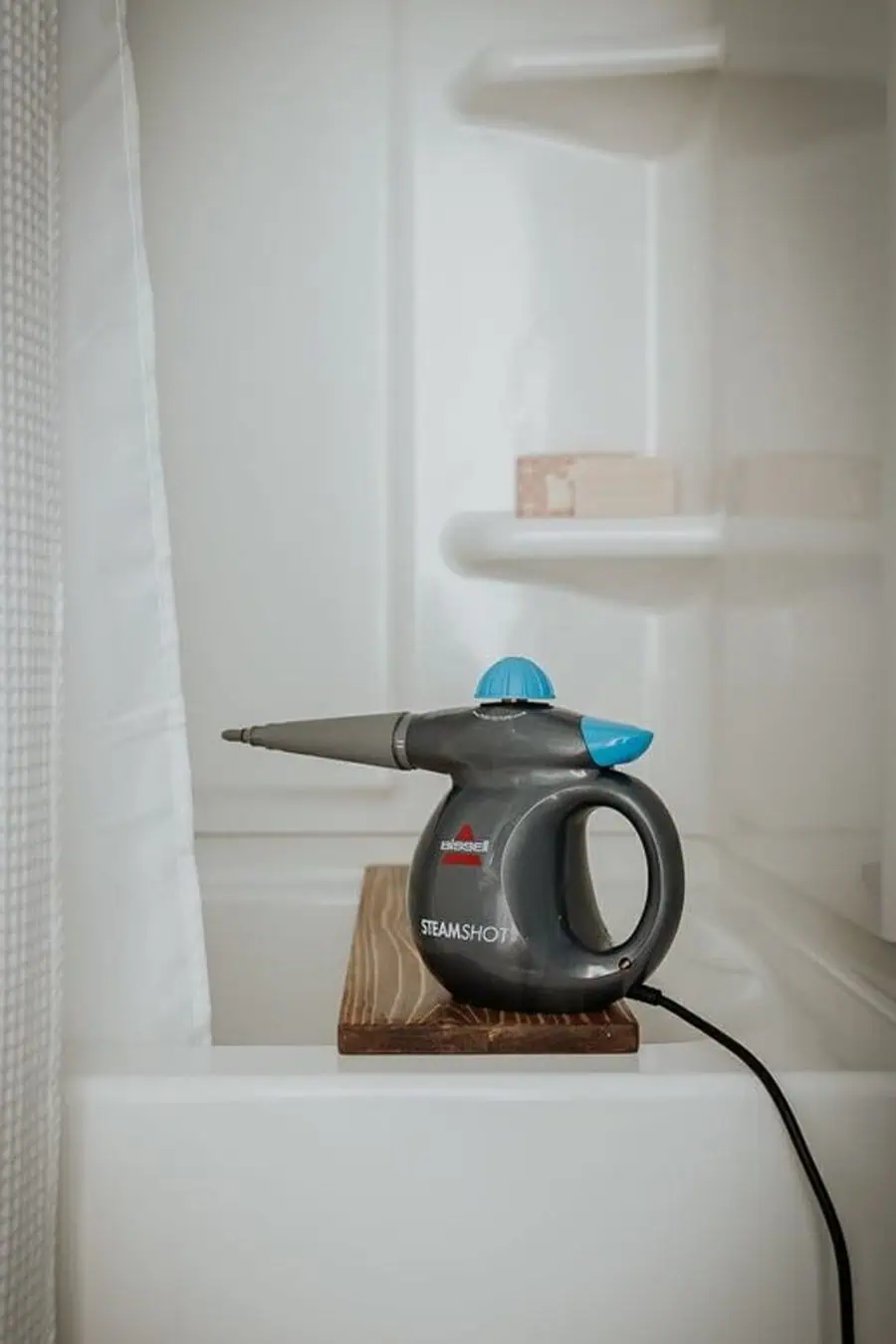 A handheld steam cleaner rests on a wooden platform in a bathtub area with a shower curtain and shelves in the background