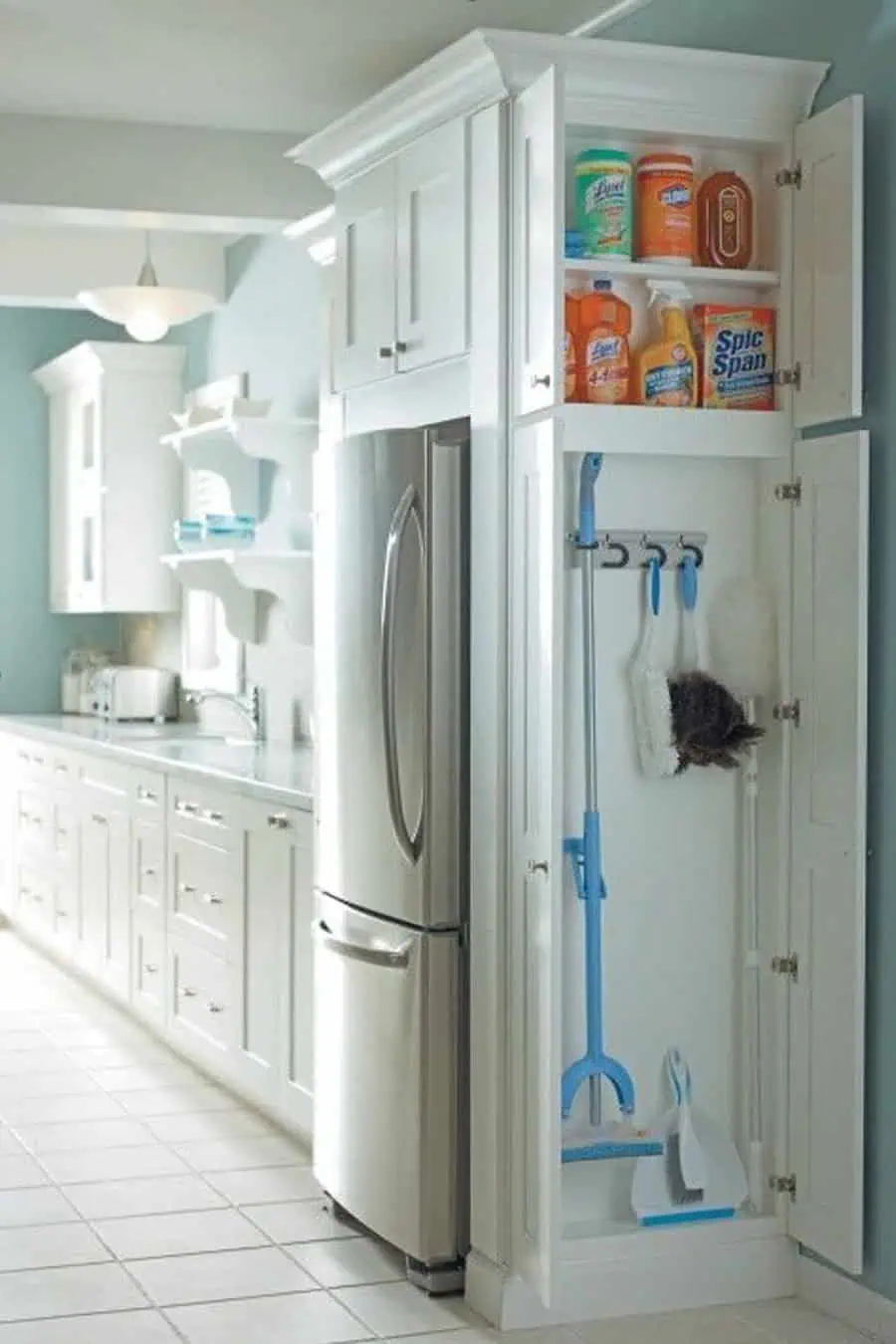 A kitchen storage cabinet with cleaning supplies and tools neatly organized next to a stainless steel refrigerator.