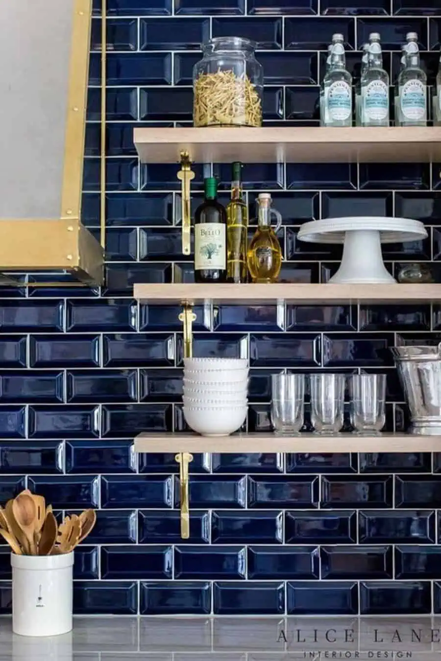 A kitchen with navy blue subway tiles, open wooden shelving, and gold hardware accents.