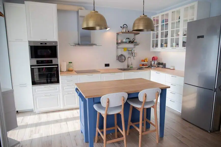 A modern kitchen with white cabinetry, a small blue island, wooden countertops, and brass pendant lights.