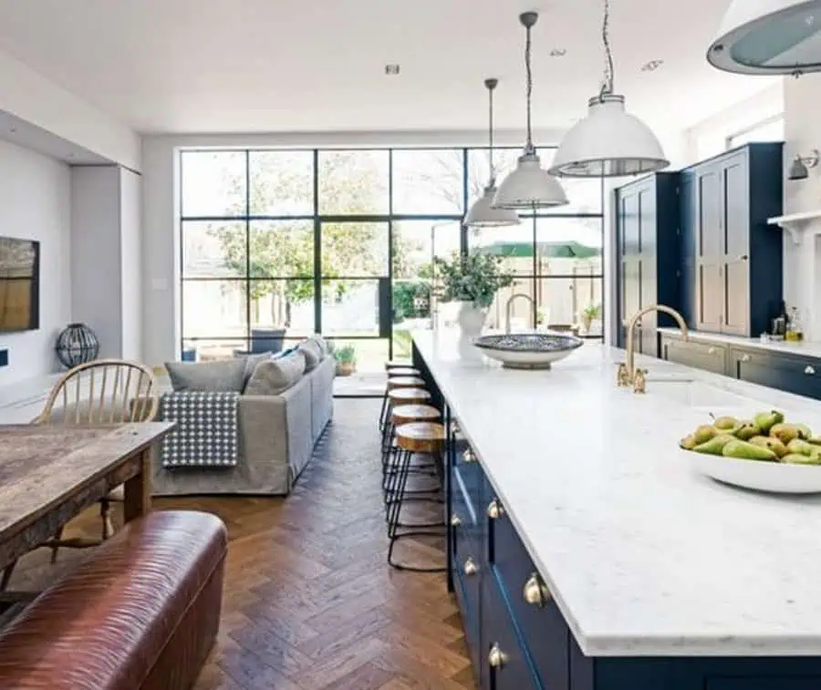 A spacious kitchen with a long marble island, navy cabinetry, and large windows overlooking the backyard, connected to a cozy living area.