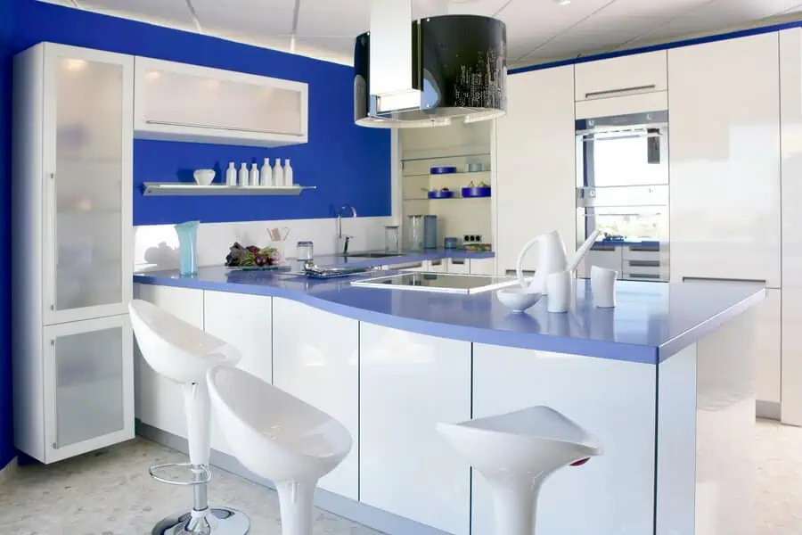 A modern kitchen with sleek white cabinets, a bright blue accent wall, and curved countertops paired with white bar stools.