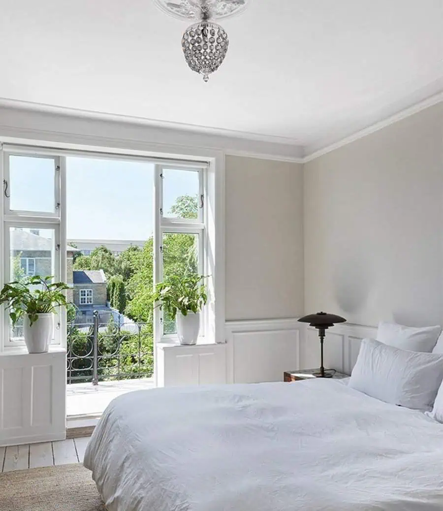 Bright bedroom with white bedding, large windows, and potted plants overlooking a garden.