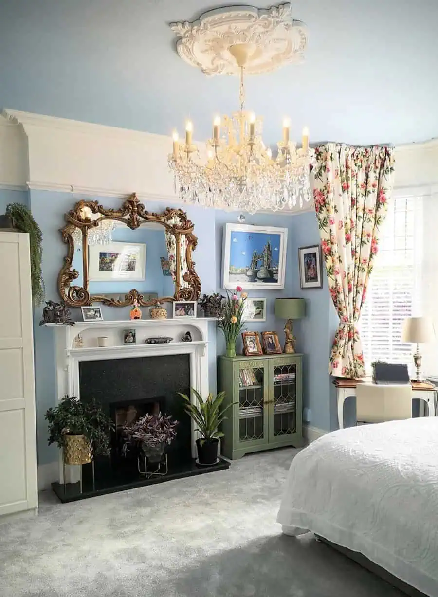 Elegant bedroom with crystal chandelier, floral curtains, and ornate gold mirror over a fireplace.