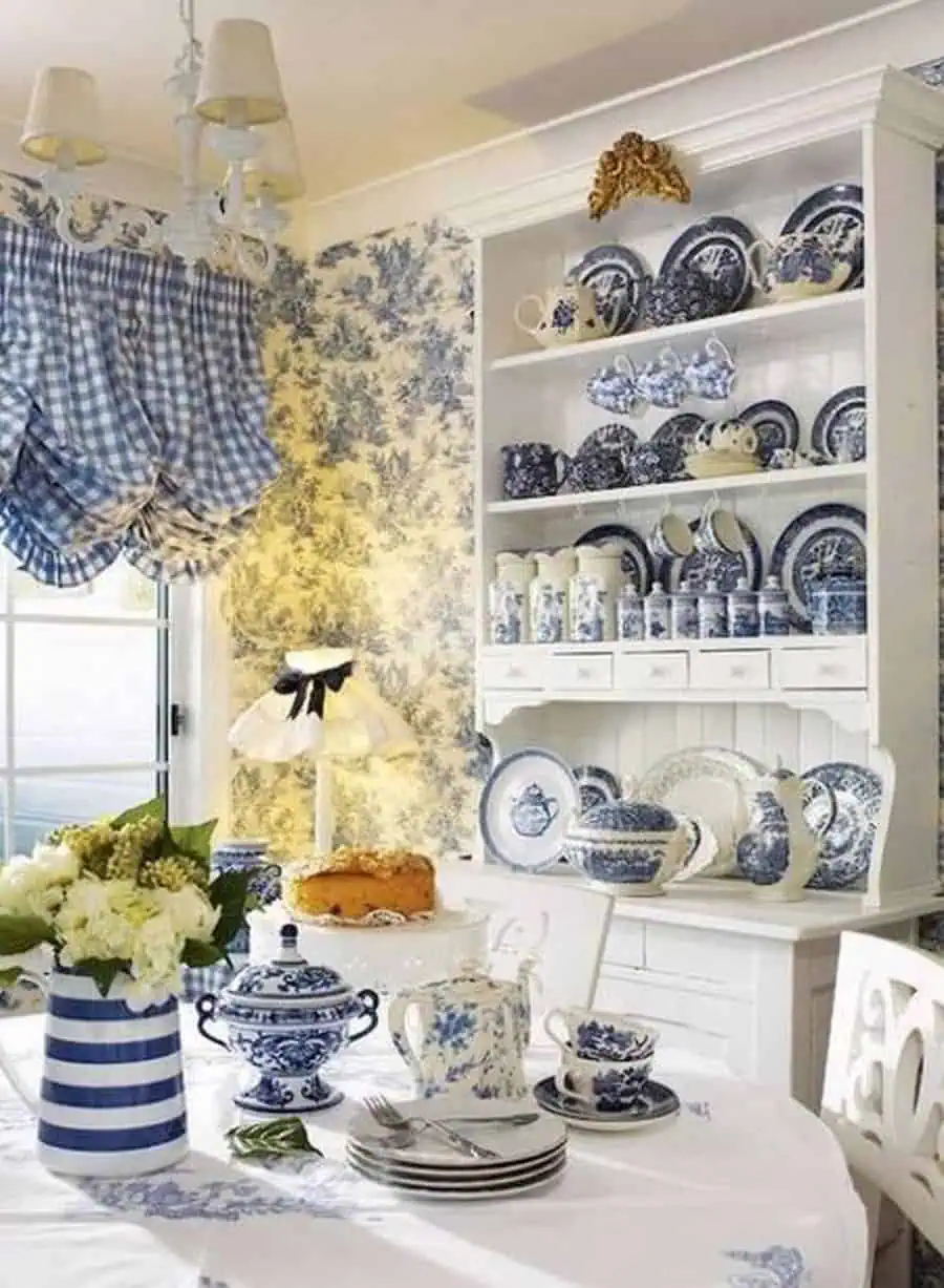 A cozy dining area with blue and white porcelain dishes displayed on a hutch, floral wallpaper, and gingham curtains.