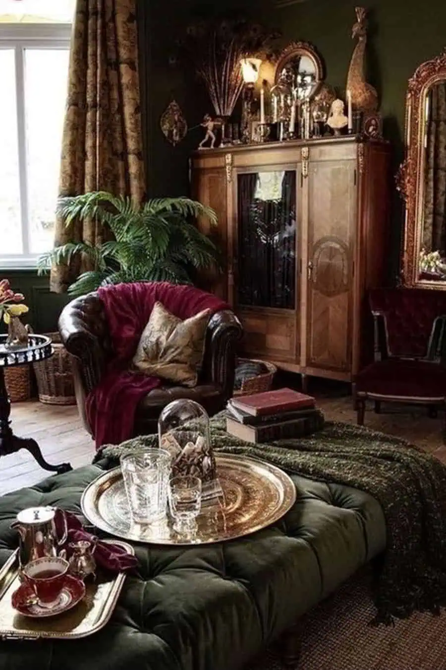 An elegant living room with a velvet green ottoman, tray of drinks, and a leather armchair. The decor includes vintage cabinets, a large mirror, and potted plants.