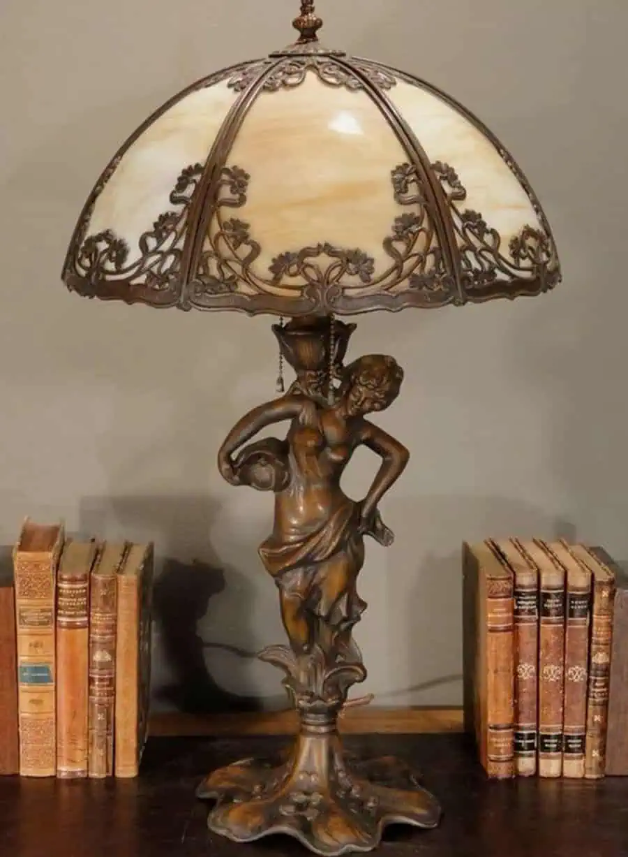 Bronze lamp with a stained glass shade, featuring a woman holding a jug, next to vintage books.