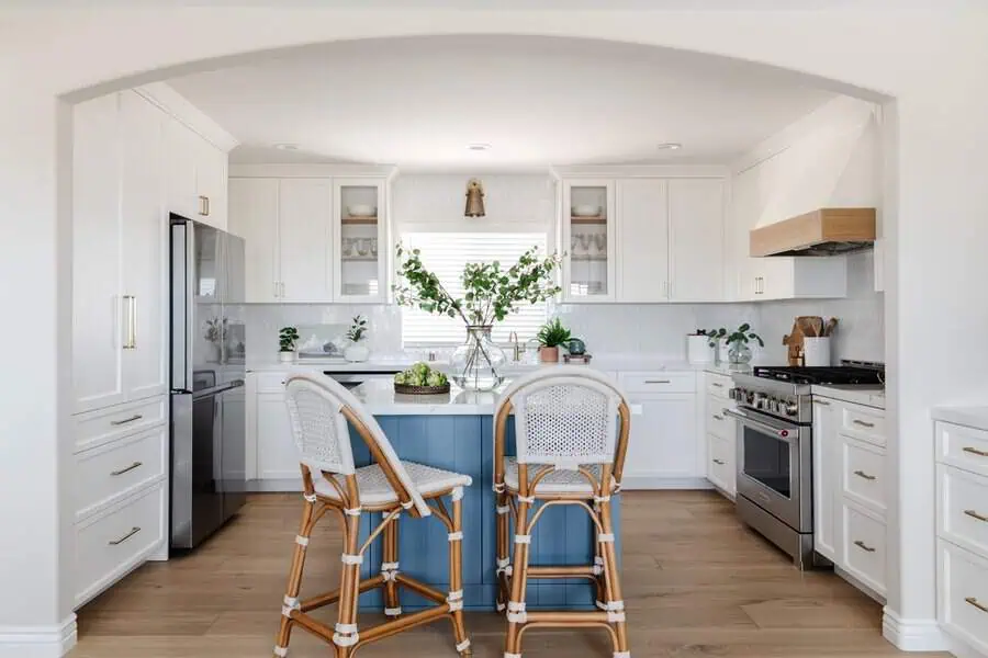 A bright and airy kitchen with white cabinetry, a blue island, and wicker bar stools under an archway.