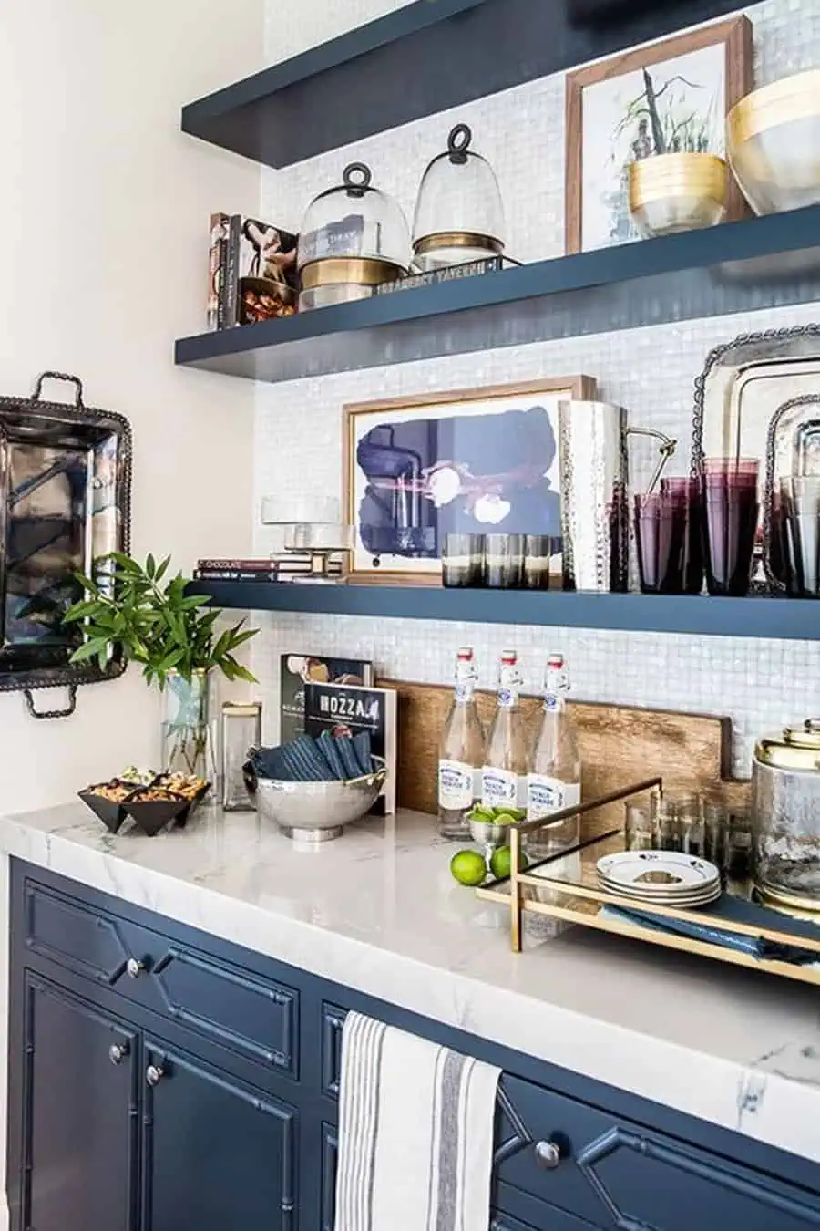 A kitchen with blue cabinets, decorative shelves, glassware, plants, and bottles on a counter