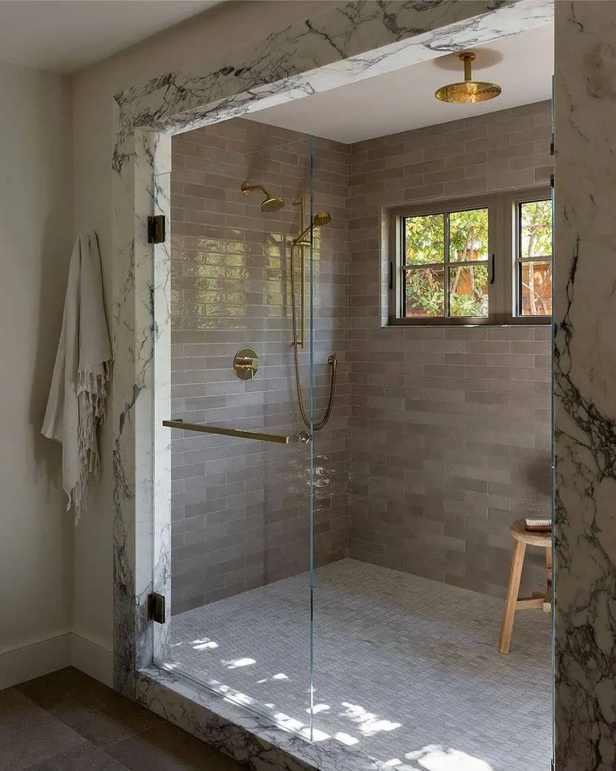 Marble-framed shower with gray subway tiles, gold fixtures, and a small window for natural light.