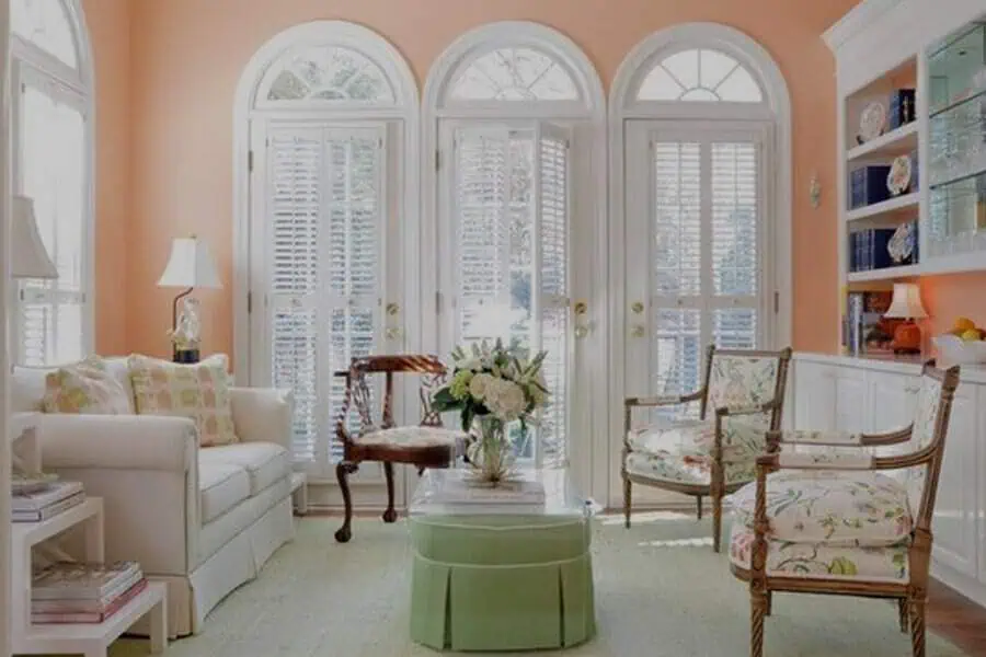 Bright sunroom with peach walls, floral chairs, white sofa, and arched French doors.