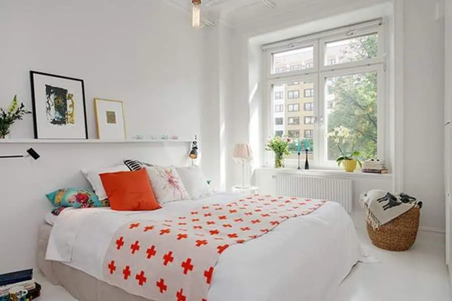 Bright bedroom with large window, white bedding, orange accents, and minimalist wall shelf decor.