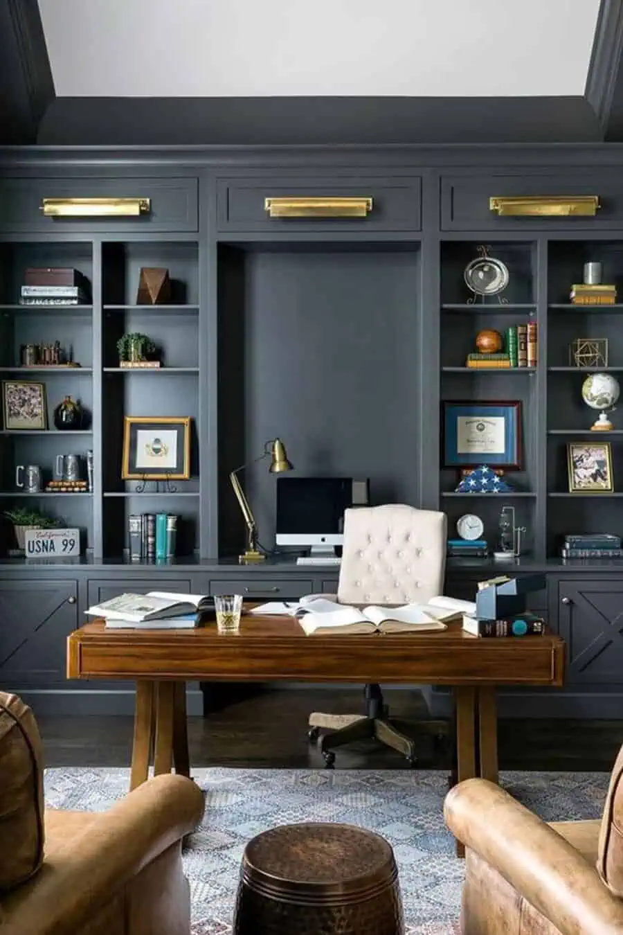 A modern home office featuring dark built-in bookshelves, a wooden desk with books and papers, and a white tufted chair.