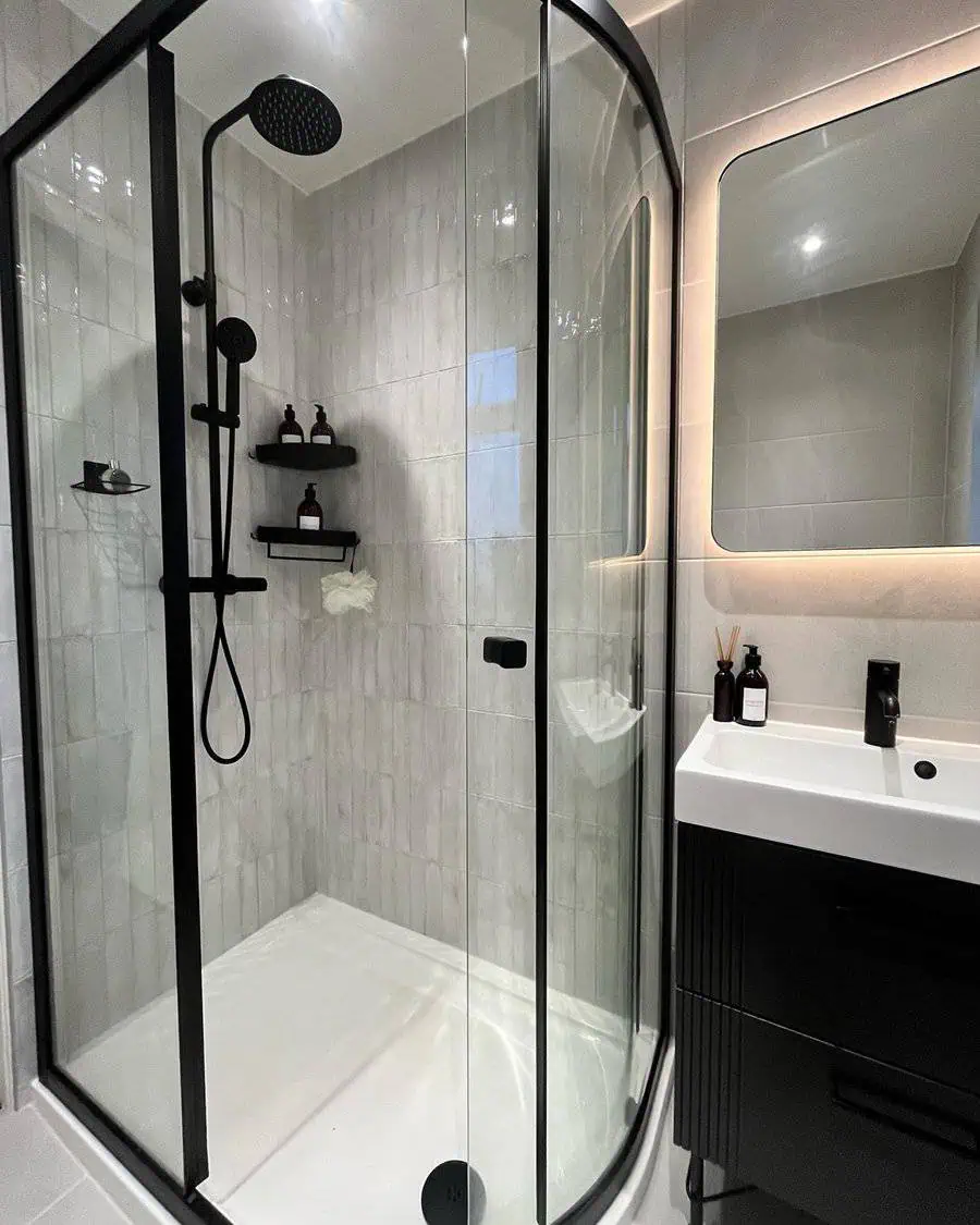 Curved glass shower with black fixtures, white tiles, and modern vanity with backlit mirror.