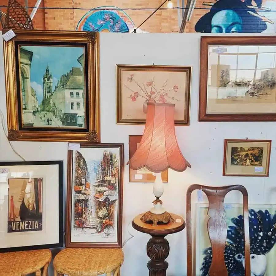 Vintage art gallery wall with framed paintings, antique lamp, and wooden chair in cozy setup.