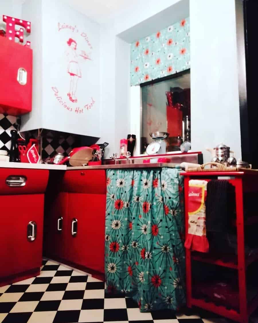 1950s kitchen with red cabinets, checkered floor, and floral-patterned curtain accents.